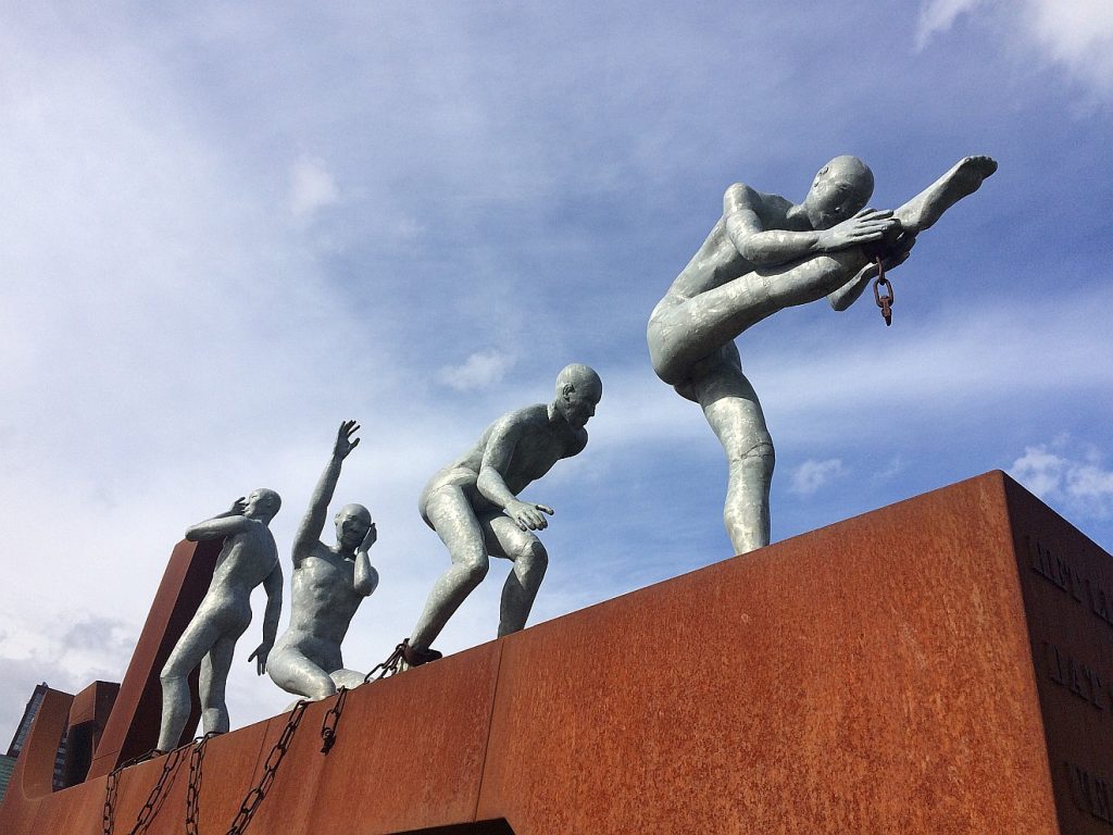 Ein Mensch ist kein Ding: Das Bild zeigt die Skulptur von vier Männern, die sich von Ketten befreien und auf einem großen rostigen Eisenträger stehen