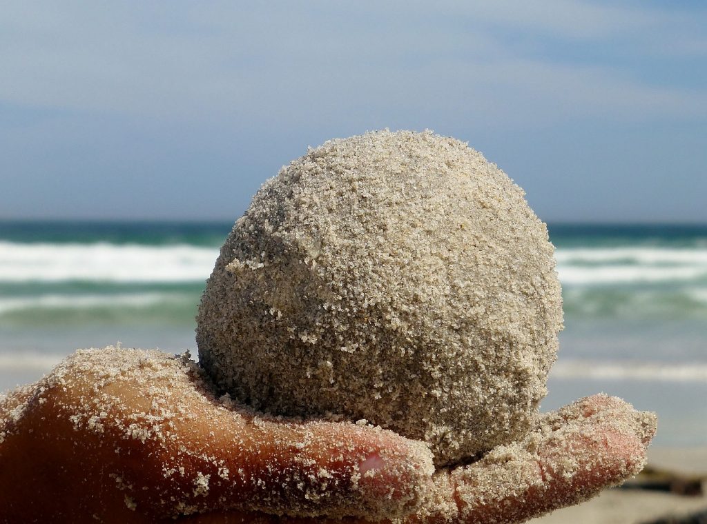Eine Handvoll mit Ruhe: Auf einer Hand liegt eine Kugel aus Sand, im Hintergrund sieht man das Meer