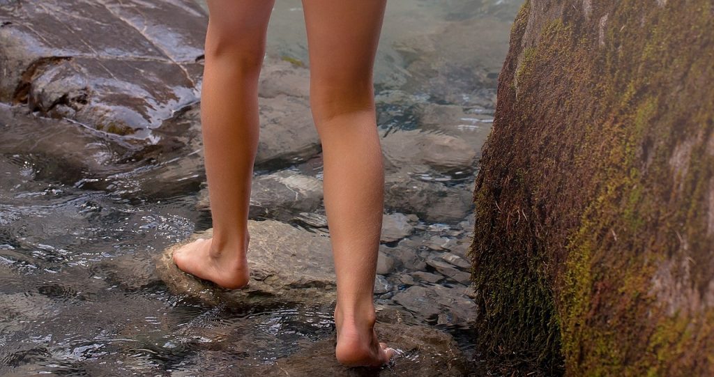 Die Beine einer Person, wahrscheinlich Frau, gehen barfuß über Steine und einen kleinen Bach