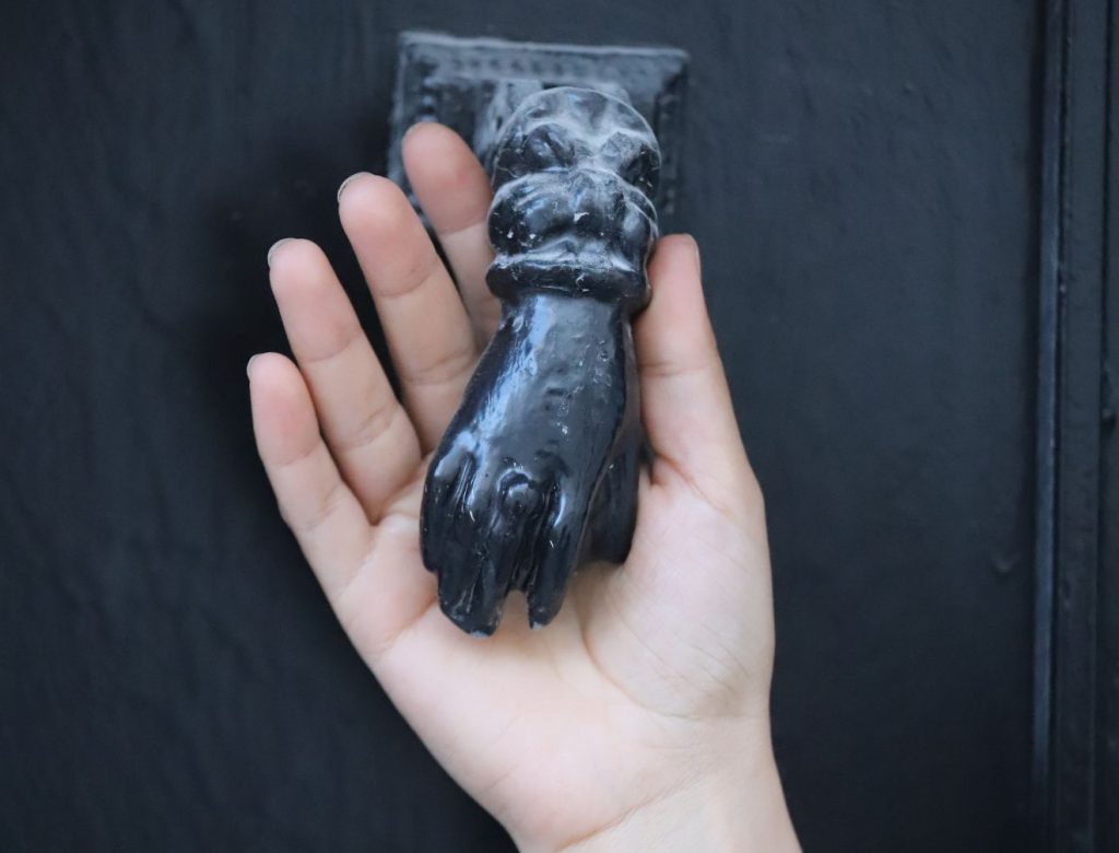 Eine schmale weiße Hand hält einen schwarzen handförmig gestalteten Türklopfer in der Hand