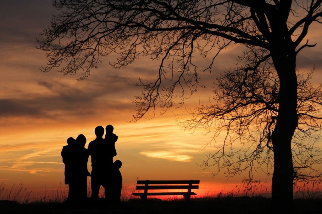 Die Silhouette eines Paares, mit zwei Kindern auf dem Arm und einem Kind an der Hand neben einer Bank und einem Bank vor einem Sonnenuntergang