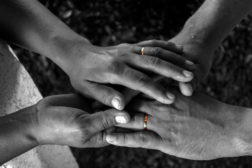 Die vier Hände eines Paares fassen sich gegenseitig - von der dunklen Farbe heben sich nur die beiden Trauringe ab