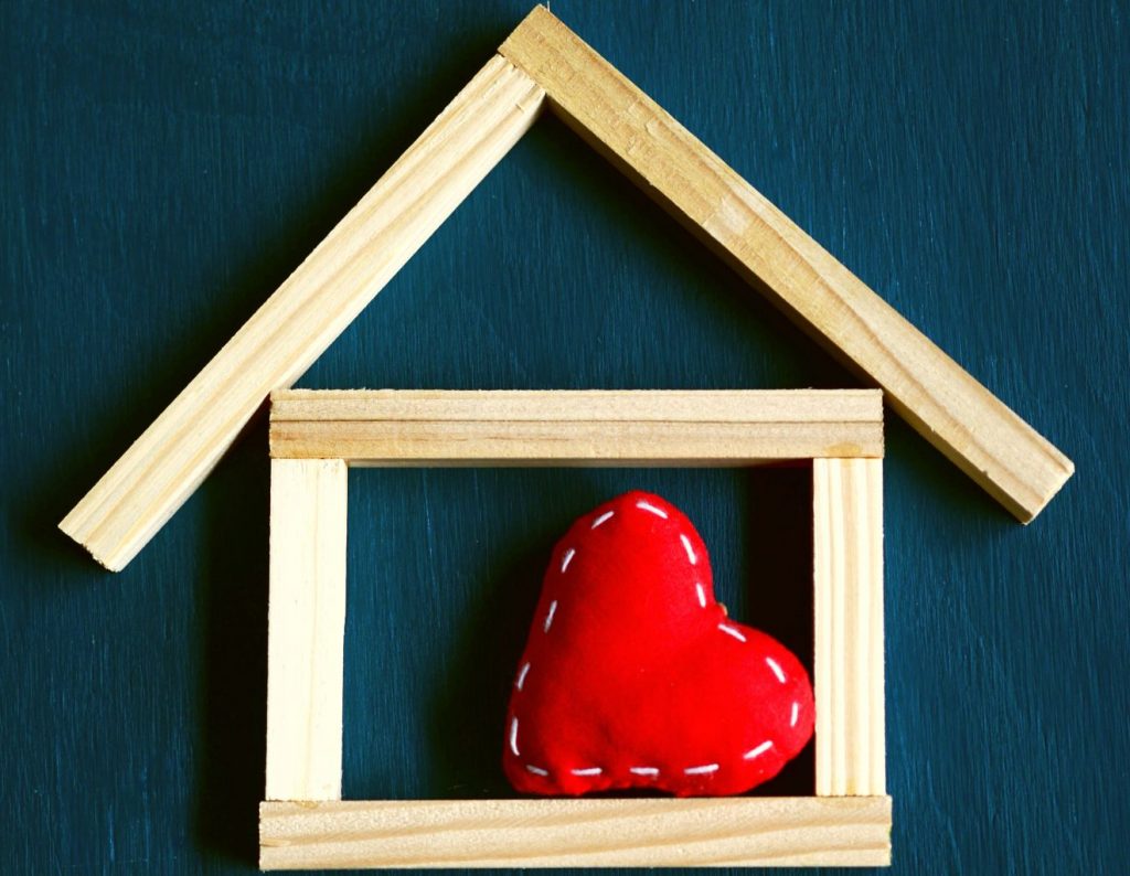 Ein stilisiertes Haus besteht aus sechs länglichen Bauklötzen, von denen vier zu einem Rechteck zusammengefügt sind, und darüber bilden zwei rechtwinklig zusammengeklebte das Dach. Im Haus liegt ein rotes Herz aus Stoff gemütlich auf einer Seite.