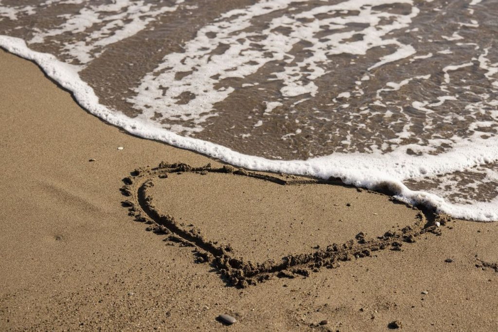 Im Sand eines Strandes ist ein Herz gemalt, das heranlaufende Wasser des Meeres droht das Herz zu überspülen.
