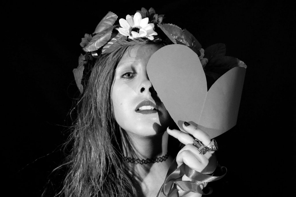 Schwarzweißbild vom Kopf einer jungen Frau vor schwarzem Hintergrund, die Blätter und eine weiße Blume im Haar trägt und mit ernstem Gesichtsausdruck sich ein aus Papier ausgeschnittenes Herz vor das linke Auge hält