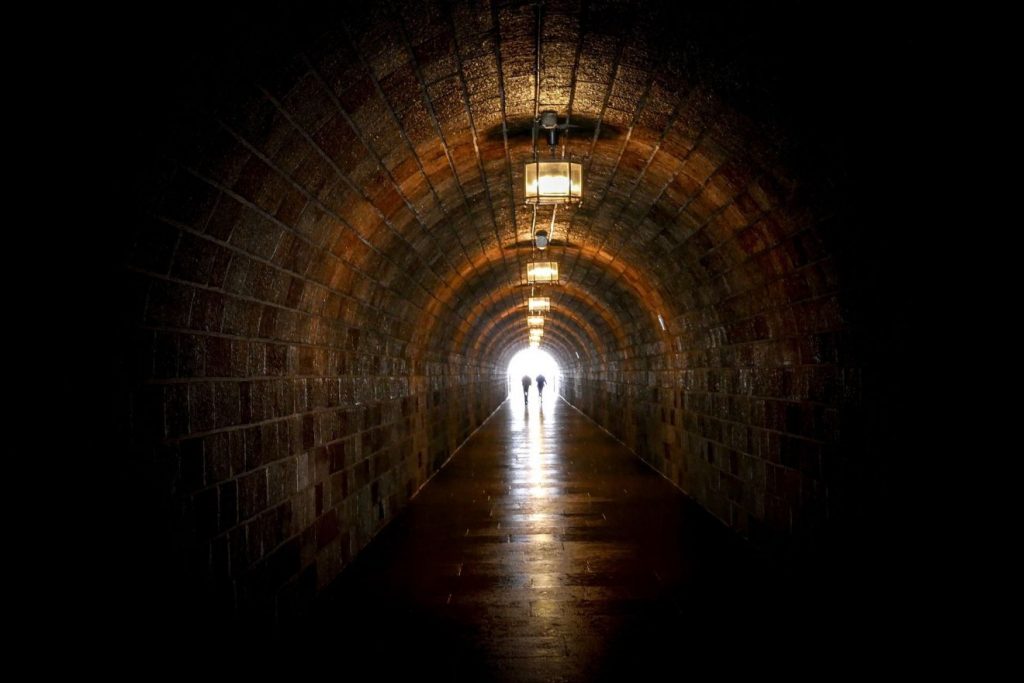 In einem dunklen funzlig erleuchteten Tunnel sind am Ende zwei Menschen zu sehen, die gerade ins helle Licht am Ende des Tunnels hineingehen.