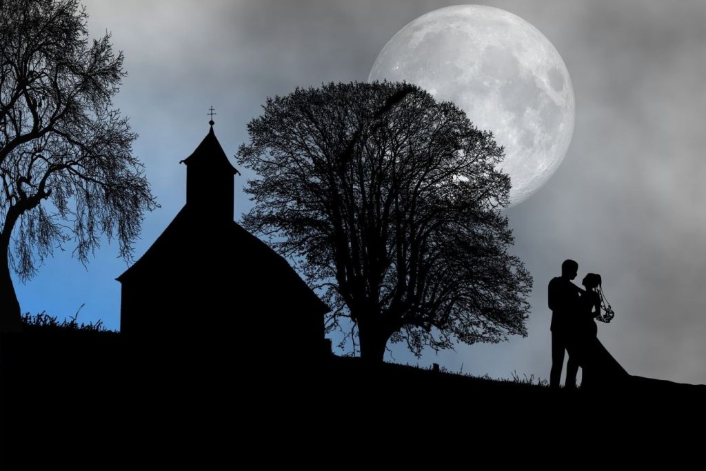 Die Silhouette eines Liebespaars, das sich anschaut, neben der Silhouette einer Kapelle, die zwischen Bäumen steht, in der Dämmerung vor einem riesigen Mond am Himmel