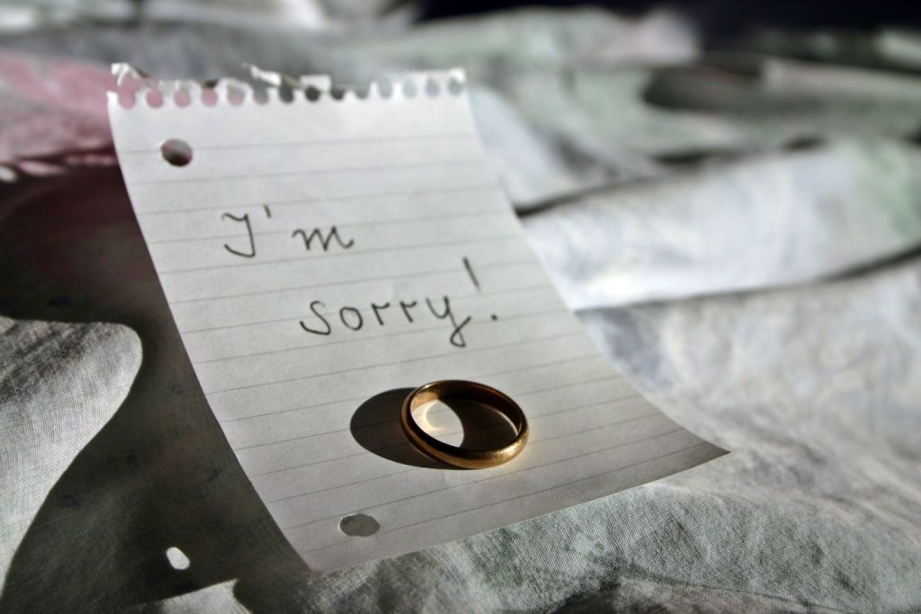 Auf einem Notizzettel, der auf einer zerknäulten Decke liegt, liegt ein Ehering mit dem Worten: "I'm sorry!" = "Es tut mir leid!"