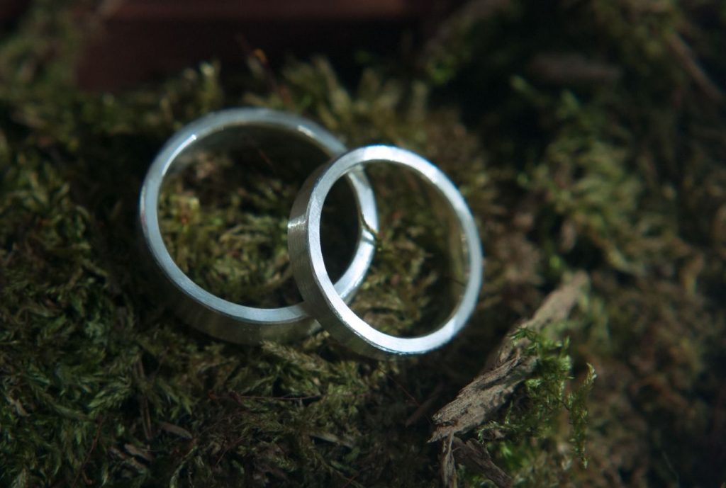 Zwei schlichte Eheringe, vermutlich aus Platin, liegen aufeinander auf einem Untergrund, der wie Waldboden aussieht