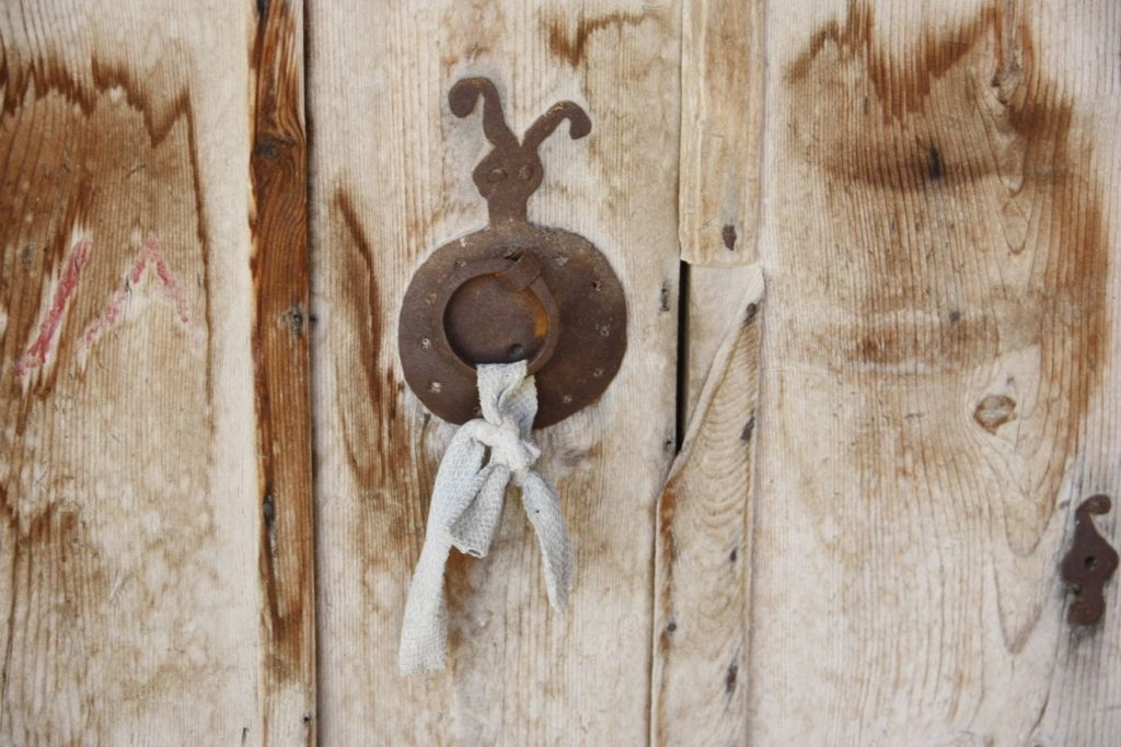Ein unscheinbarer Türklopfer, der nur aus einem verrosteten Ring mit einem daran geknoteten Stück Stoff an einem rostigen Schild besteht, ist mit Nägeln an einer uralten Holztür befestigt.