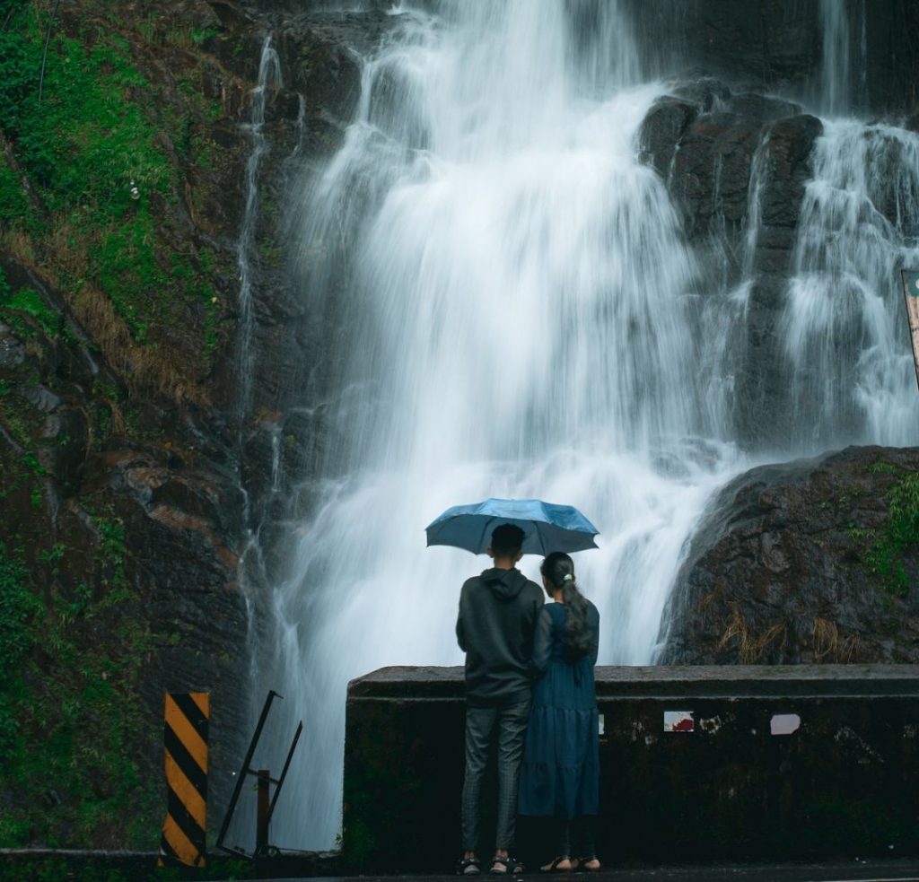Ein Liebespaar steht mit einem Regenschirm vor einem Wasserfall.