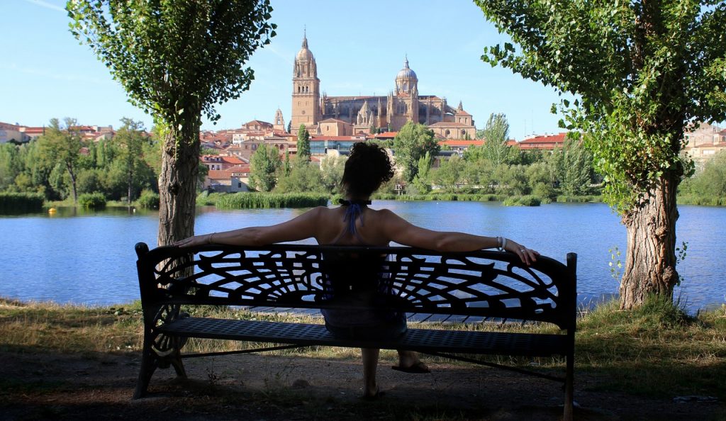 Eine Frau sitzt mit dem Rücken zum Betrachter mit ausgebreiteten Armen auf einer Parkbank vor einem Fluss und zwei Bäumen, hinter denen eine große Kirche zu sehen ist