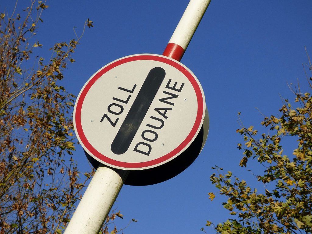 Eine Zollschranke mit dem Schild "Zoll / Douane" erhebt sich in den blauen Himmel