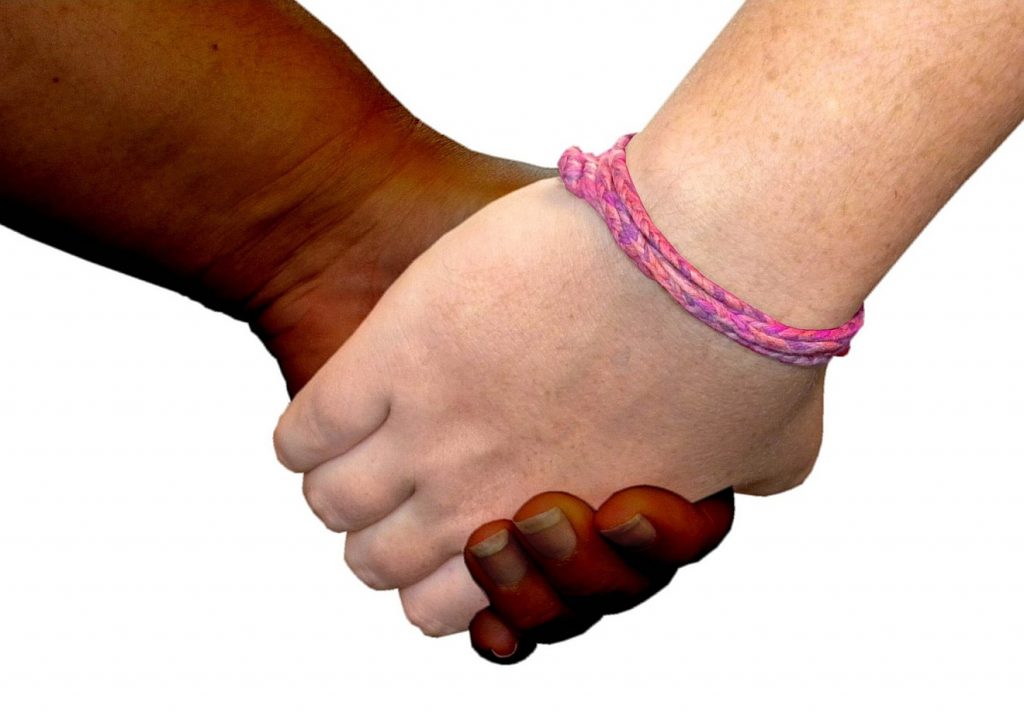 Unkonventioneller Glaube und der verletzbare Gott: Zwei Hände unterschiedlicher Hautfarbe halten einander, eine davon mit rosa Bändchen