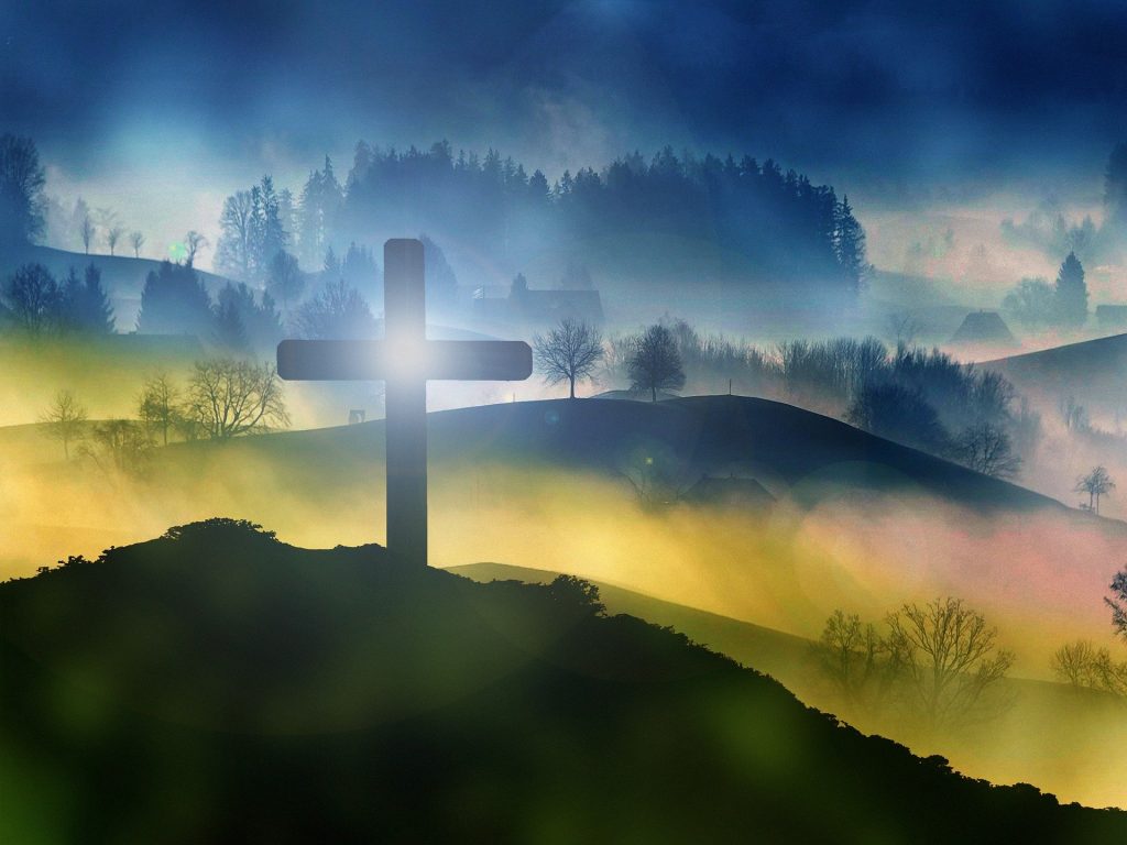 Eine hügelige Landschaft, vorn und hinten dunkel, in der Mitte heller, mit Nebelschwaden dazwischen. auf dem dunklen Hügel vorne steht ein schwarzes Kreuz