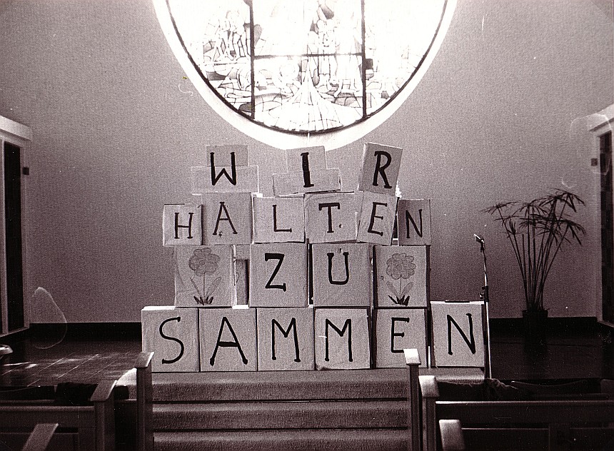 Foto von einem Familiengottesdienst 1983 mit einer Mauer aus Kartons mit dem Schriftzug: "Wir halten zusammen"